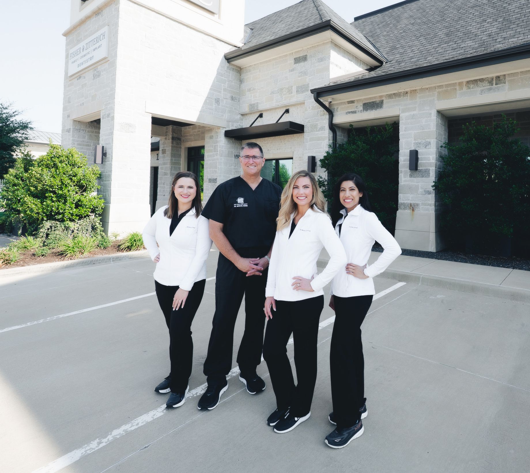 Four dental professionals standing outside a clinic with stone exterior, three women in white jackets and one man in black scrubs.
