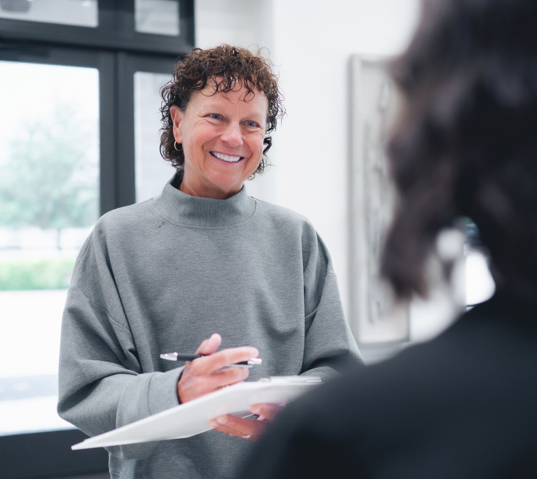 Smiling woman in gray sweater holding a clipboard and pen, engaging with another person.