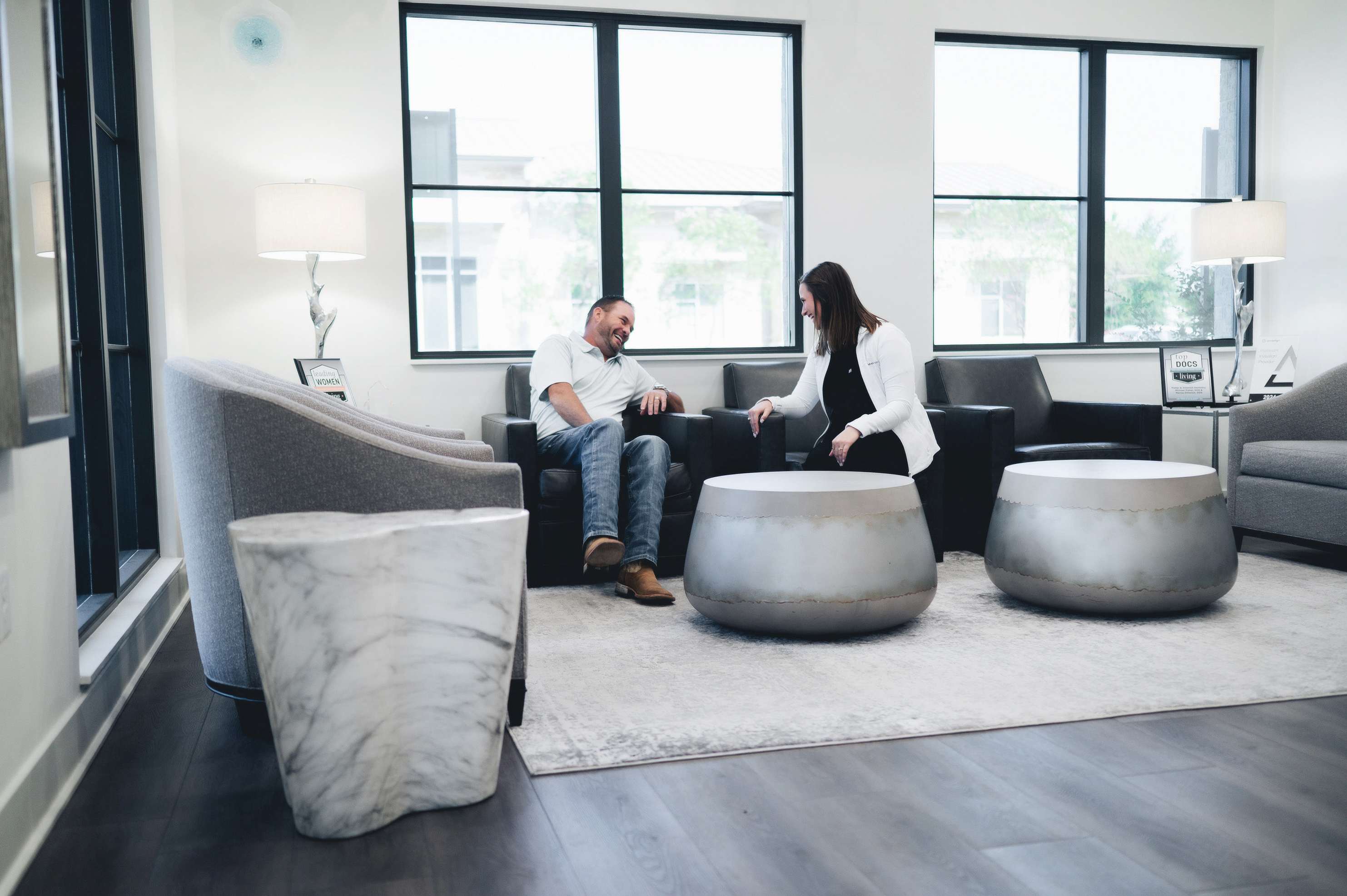 Man and woman sitting on black chairs in a bright modern living room, smiling and talking across round silver coffee tables.
