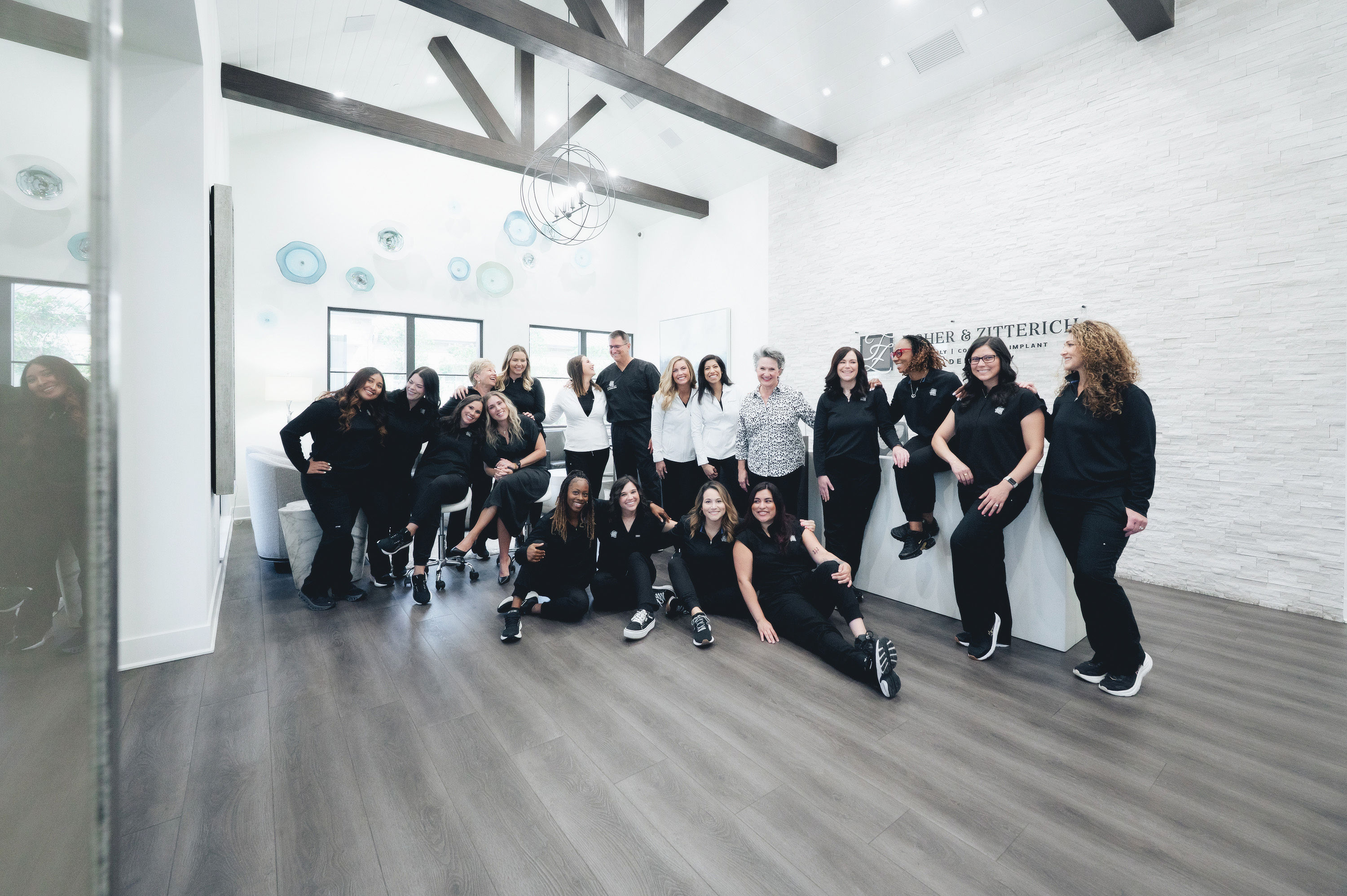 Group of dental office staff posing and smiling together in a modern reception area with wooden beams and light walls.