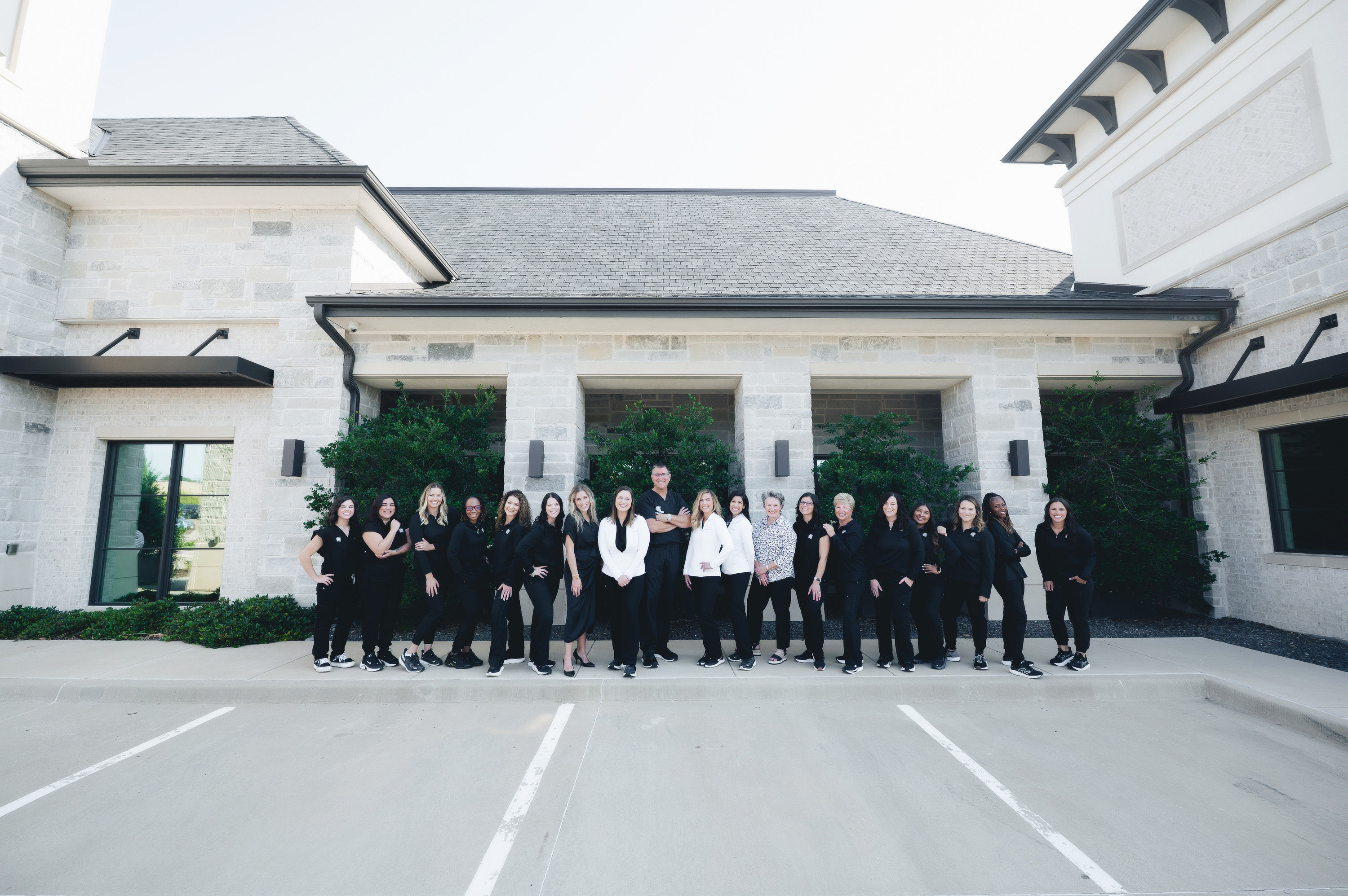 Group of 19 people standing side by side outside a modern building with stone walls and greenery, most wearing black with some in white.