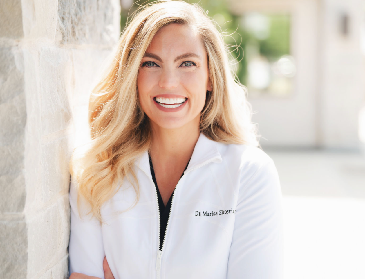 Smiling woman with long blonde hair wearing a white jacket standing against a stone wall outdoors.