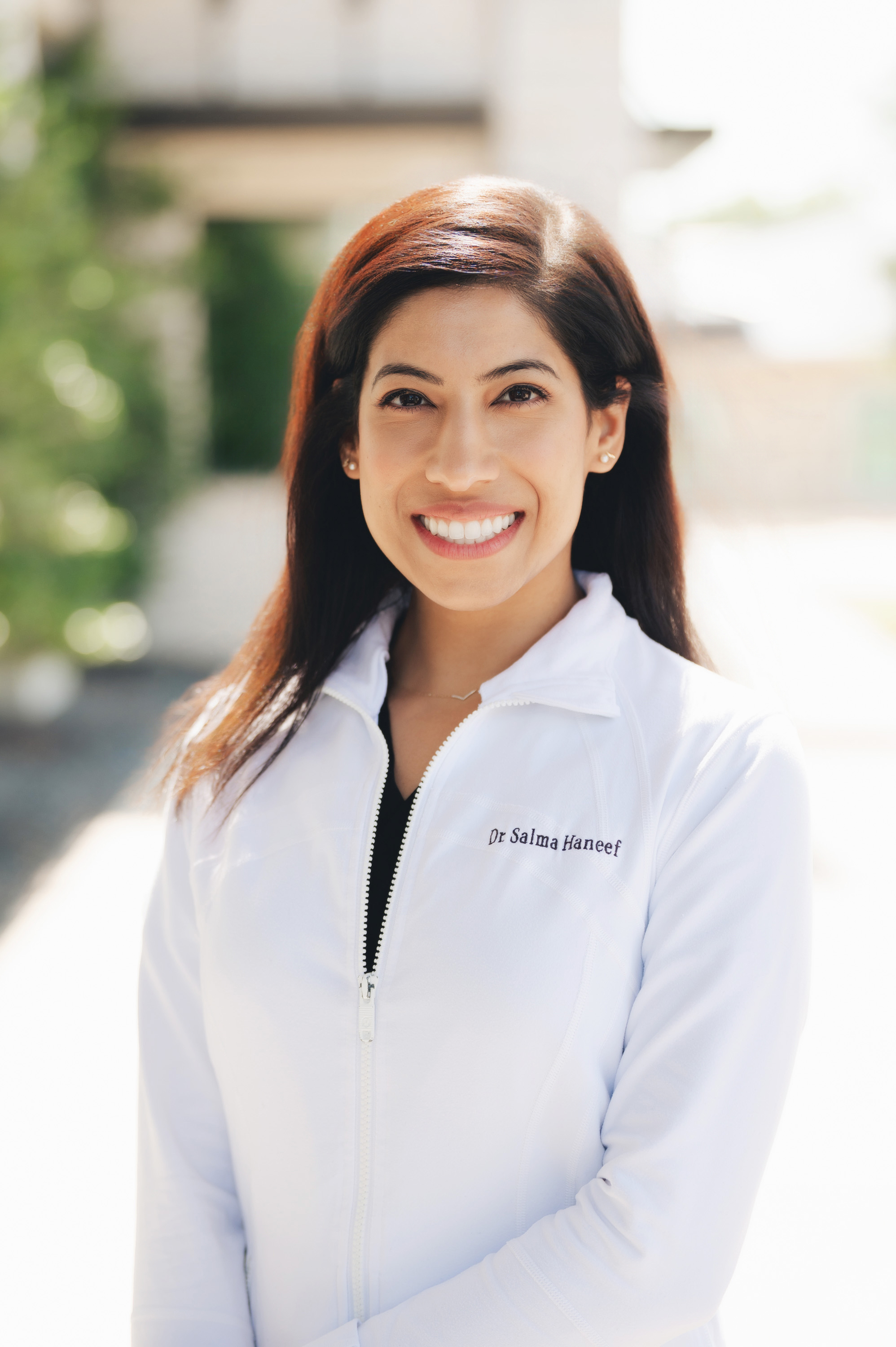 Smiling woman with long dark hair wearing a white zip-up jacket embroidered with 'Dr Salma Haneef'.