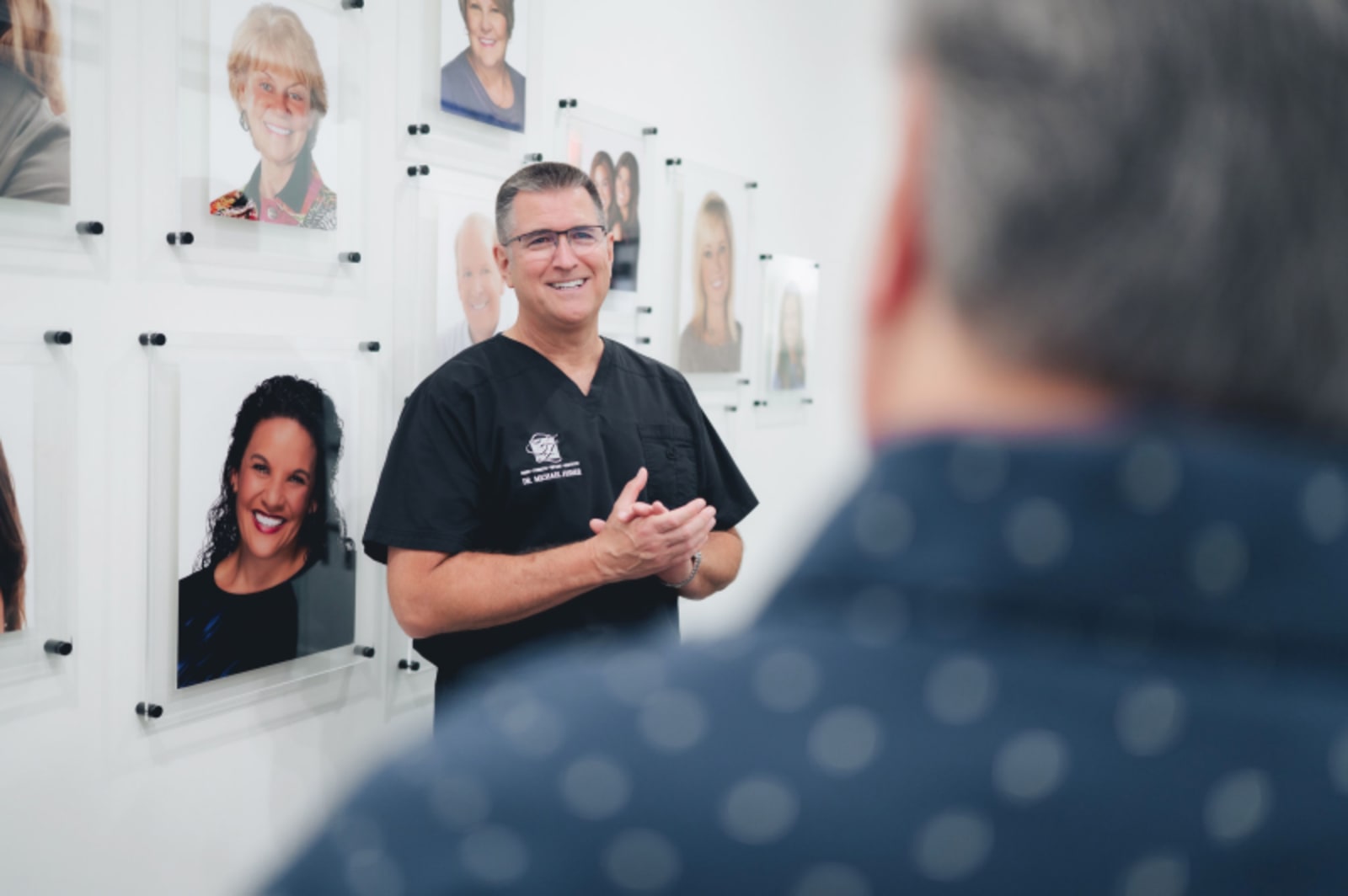 Dentist speaking with patient in modern office setting with team photos on wall behind him.