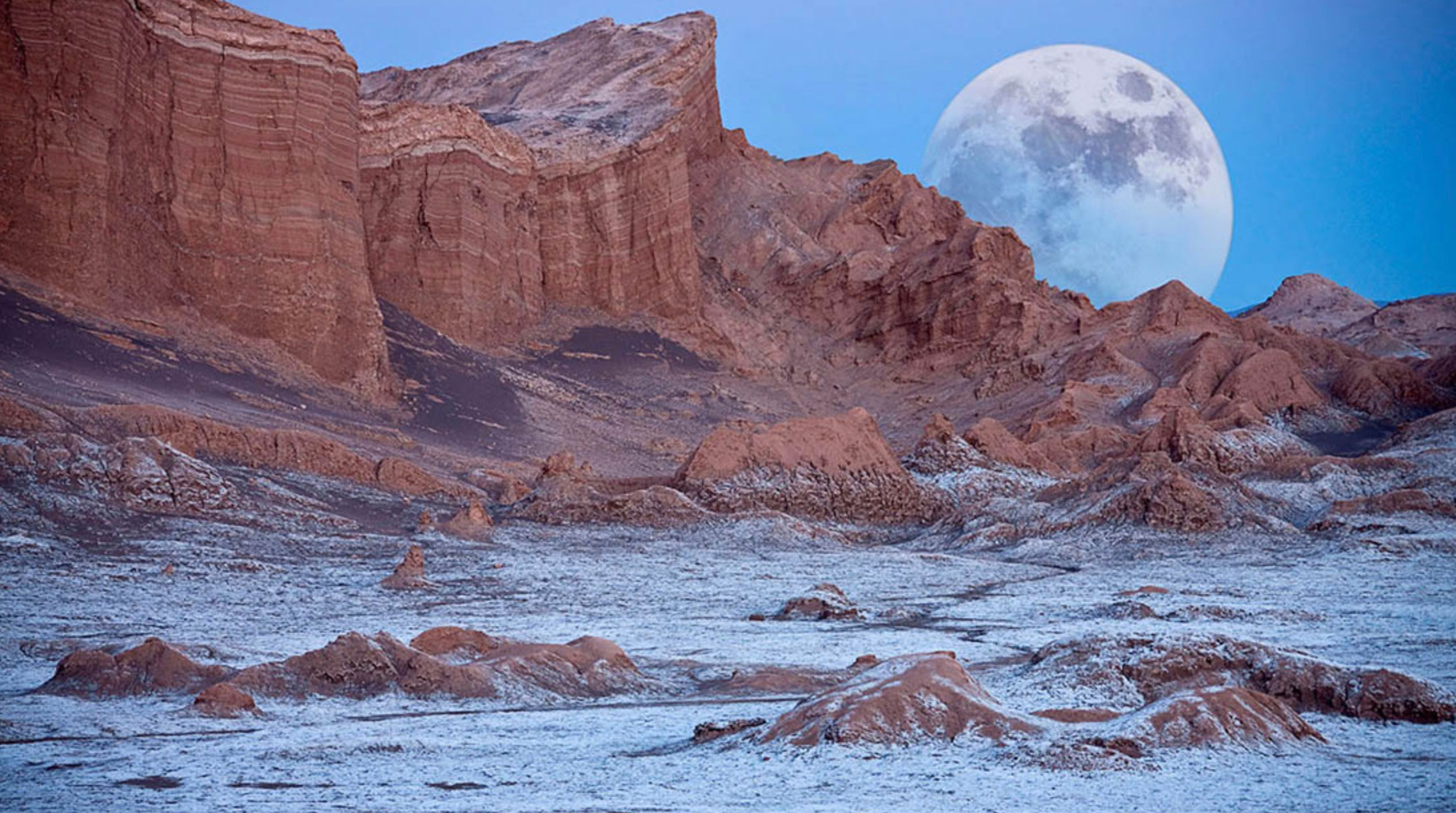 Rocky desert landscape with dusted snow and a large full moon rising behind cliffs at dusk.