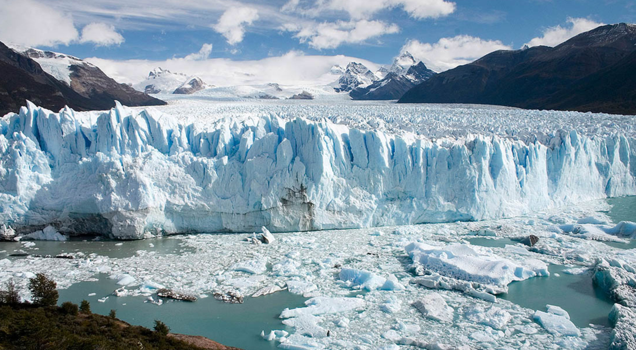 Expansive blue glacier with jagged ice formations, floating ice chunks in cold water, and snow-capped mountains under a partly cloudy sky.