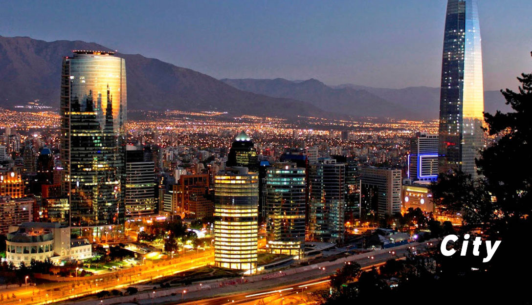 Nighttime cityscape with illuminated skyscrapers, roads, and mountains in the background.