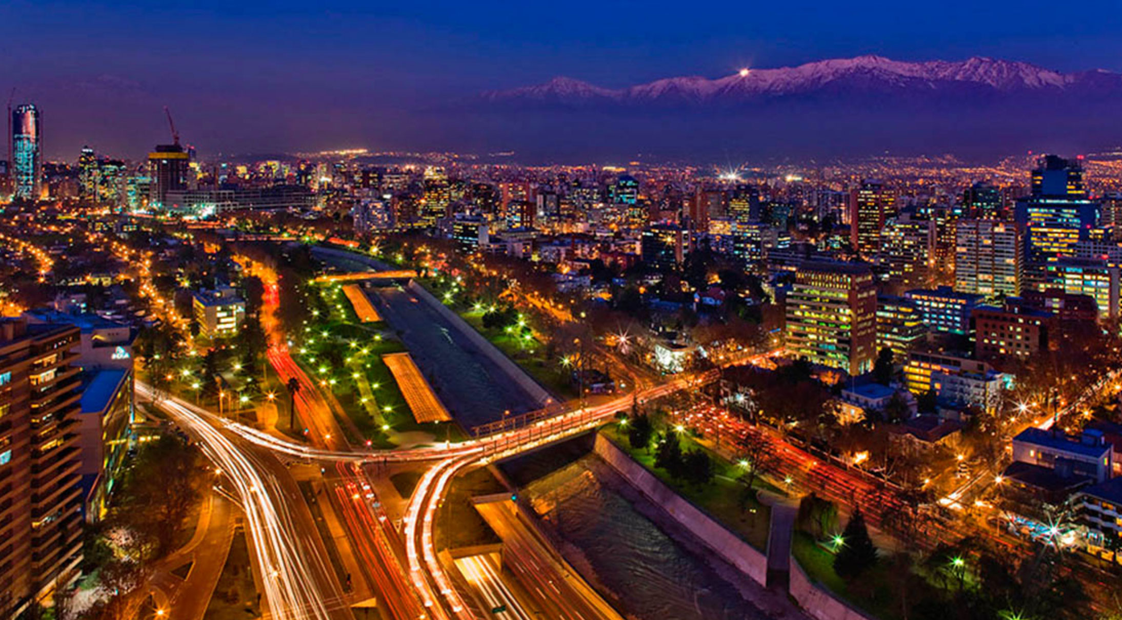 Nighttime aerial view of a city with illuminated streets, tall buildings, a river, and mountains in the background.