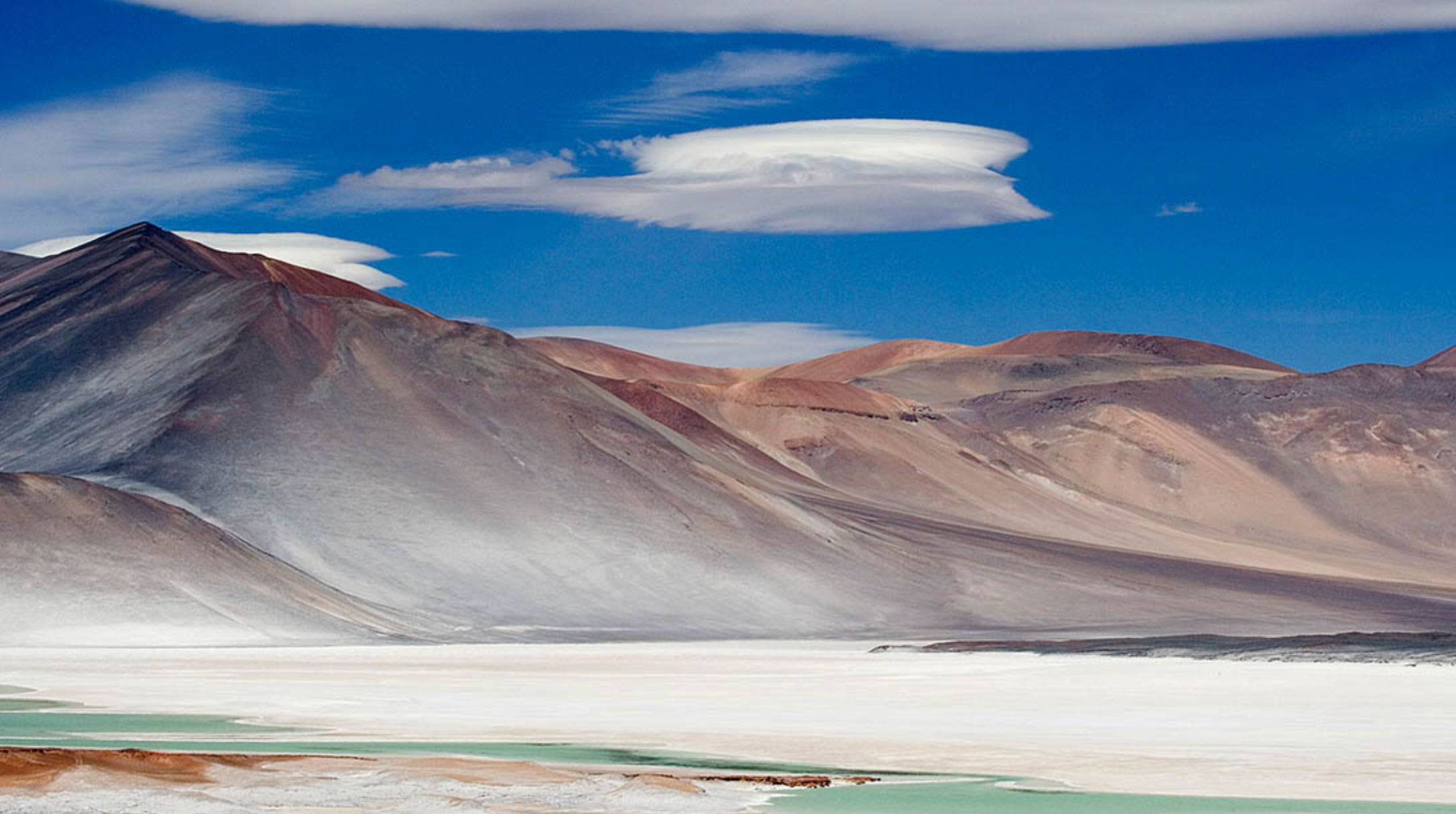 Arid mountains with reddish and gray slopes under a deep blue sky with distinct lenticular clouds above a white salt flat and patches of green water in the foreground.