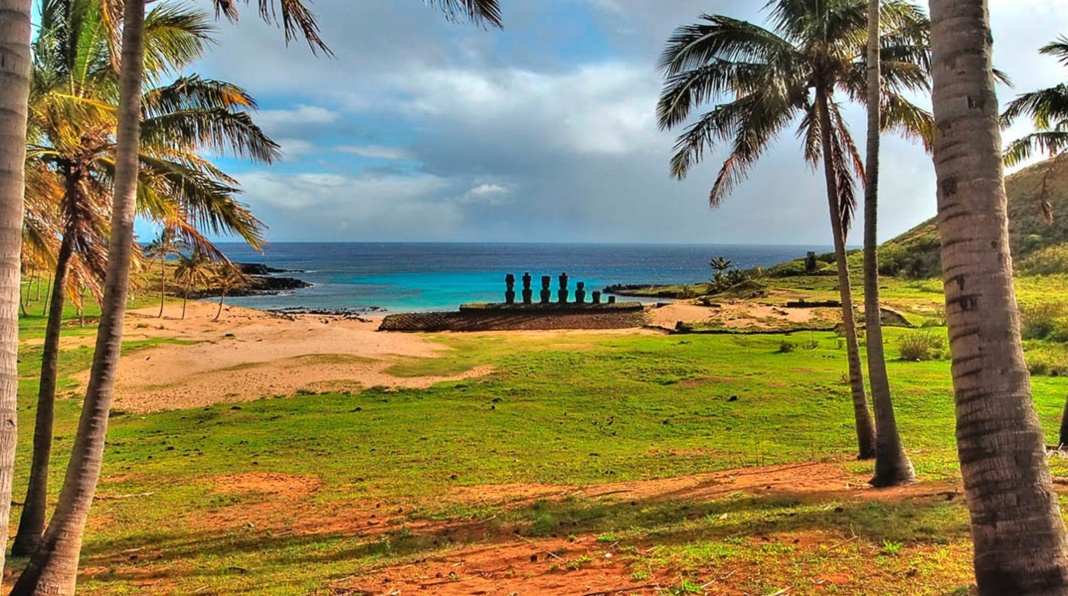 Moai statues on a beach with palm trees, green grass, and blue ocean under a cloudy sky.