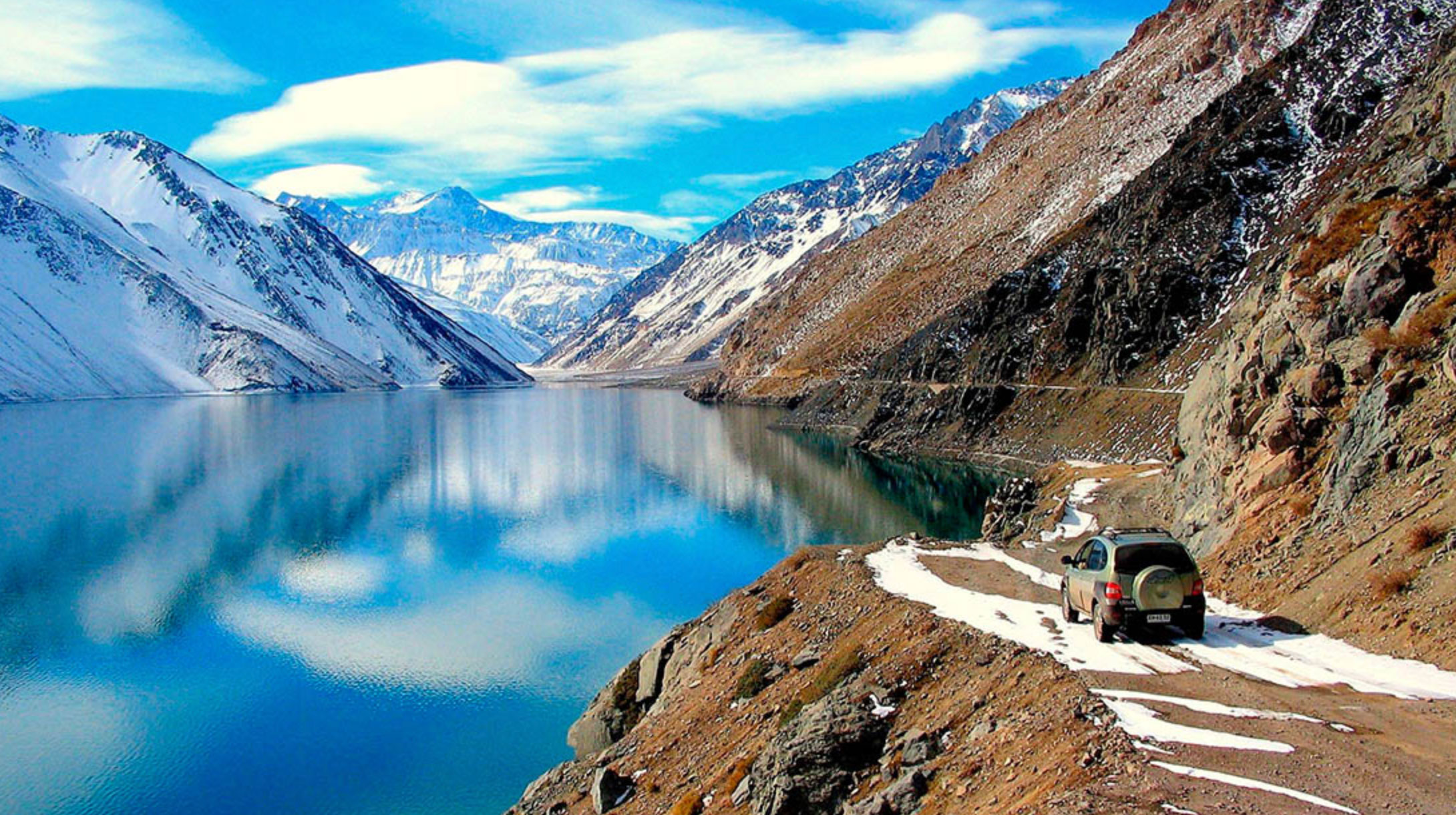 SUV driving on a snowy mountain road beside a reflective blue lake surrounded by snow-capped peaks under a partly cloudy sky.