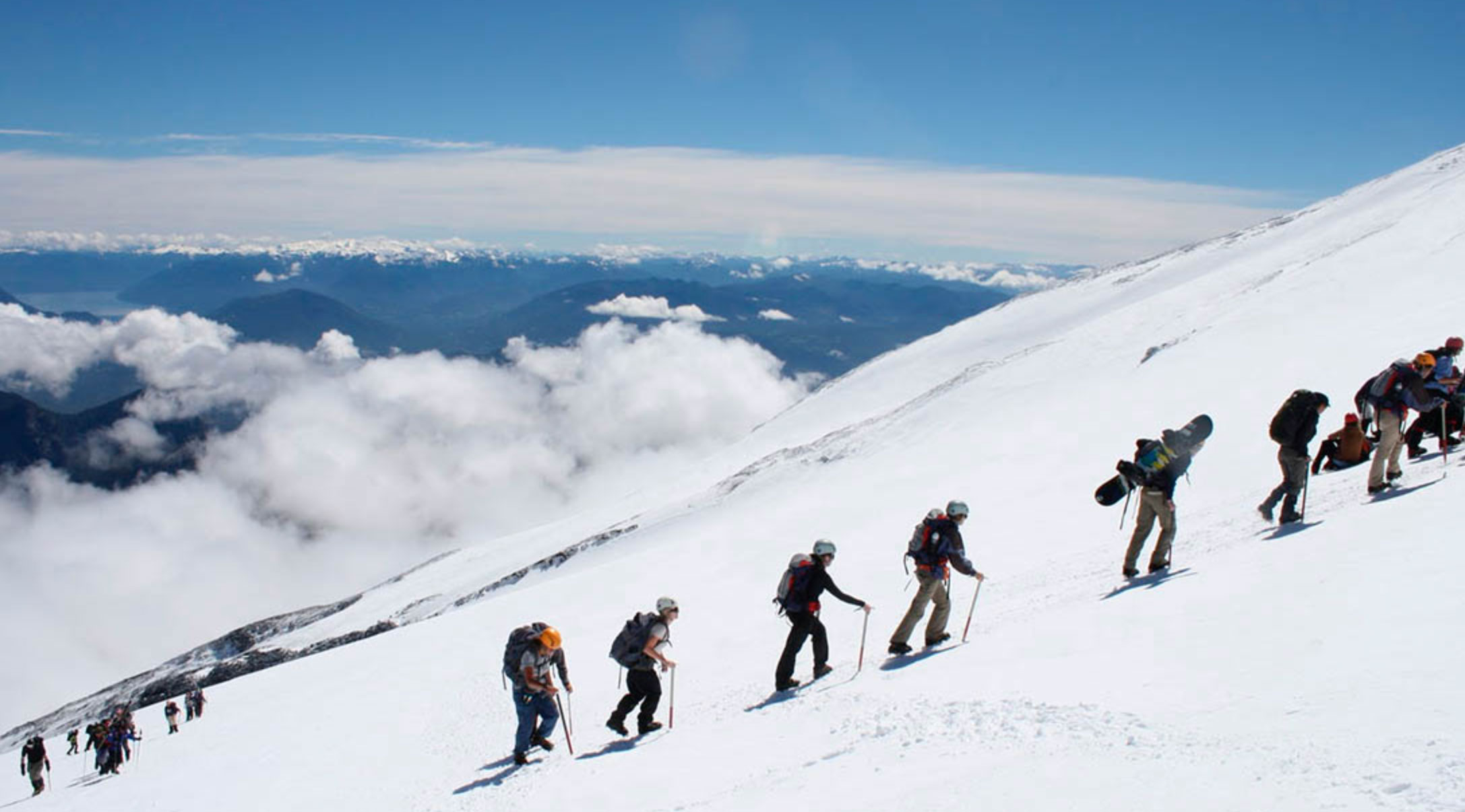 Group of climbers ascending a snowy mountain slope with mountains and clouds in the background.