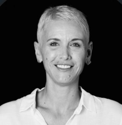 Portrait en noir et blanc d'une femme souriante aux cheveux courts et clairs, portant une chemise blanche, sur fond noir.