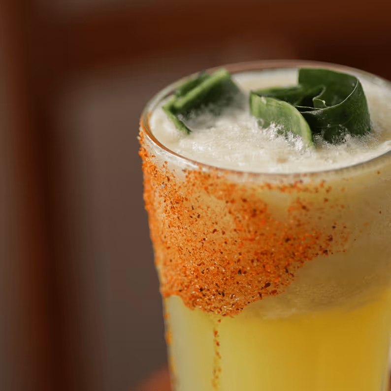 Close-up of a frothy yellow cocktail with a chili powder rim and green garnish on top.