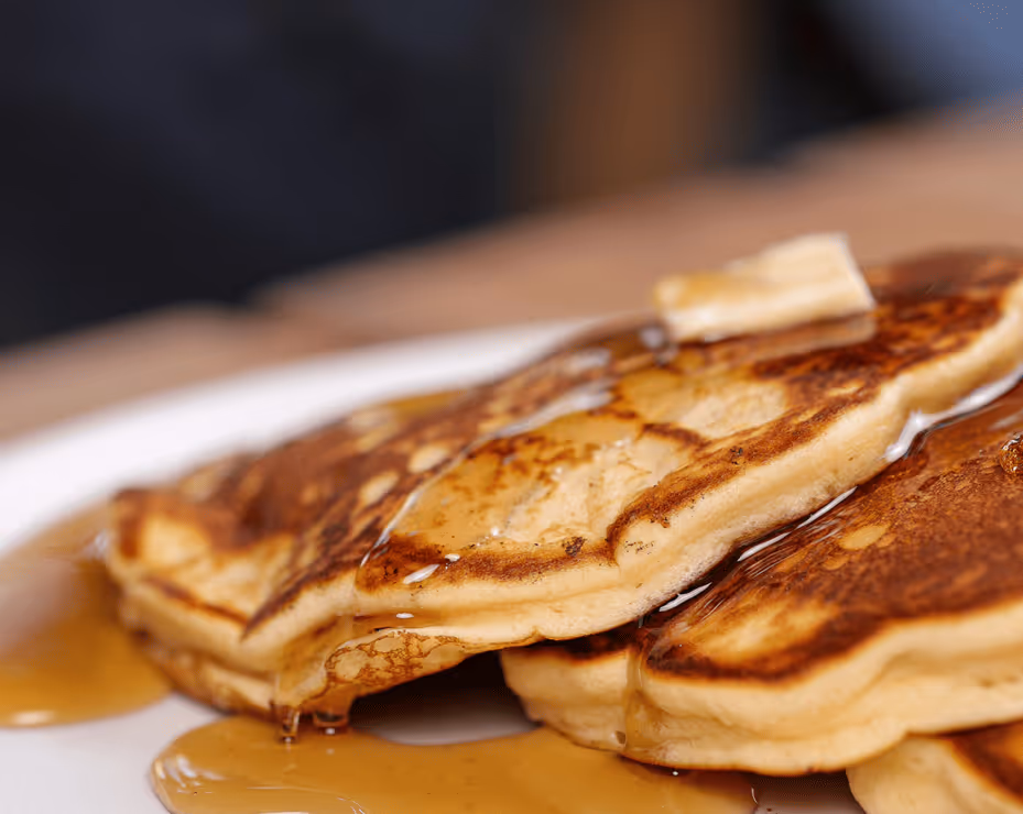 Close-up of stacked golden-brown pancakes topped with melting butter and dripping maple syrup on a white plate.