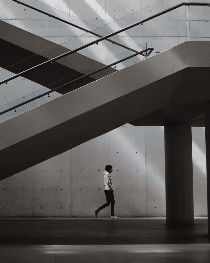 Person walking beneath modern angular staircases inside a building with concrete walls and dramatic light and shadow.