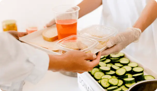 Mãos com luvas brancas entregando uma bandeja com uma fatia de pão, copo com bebida laranja e dois recipientes plásticos com comida para outra pessoa.
