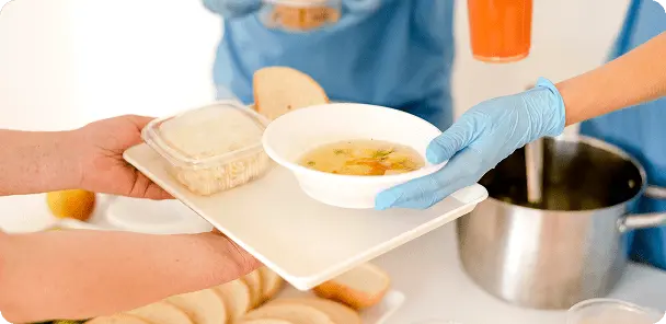 Pessoa entregando bandeja com sopa em tigela branca e recipiente plástico com comida para outra pessoa com luva azul, com panela ao fundo.