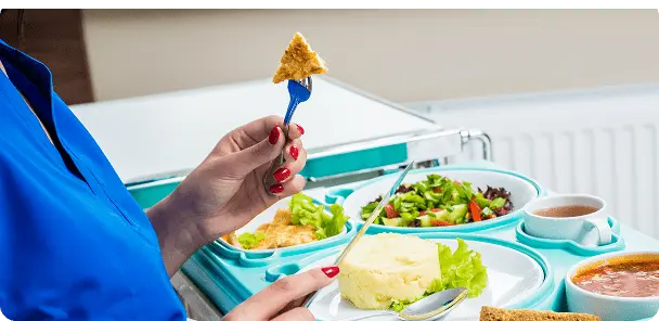 Pessoa vestindo azul segurando um garfo com um pedaço de tortilha sobre um prato com purê, salada e sopa em bandeja hospitalar.