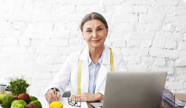 Nutricionista sorrindo, com bata branca e fita métrica amarela no pescoço, sentada à mesa com laptop, caneta e verduras em jarros, parede de tijolos branca ao fundo.