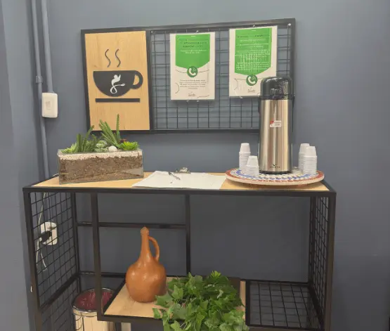 Mesa com cafeteira, copos, plantas e certificados pendurados em parede azul.