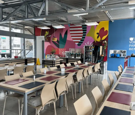 Refeitório moderno com cadeiras bege e mesas organizadas com jogos americanos vermelho e preto, parede colorida com arte abstrata e uma estação de bebidas ao fundo.