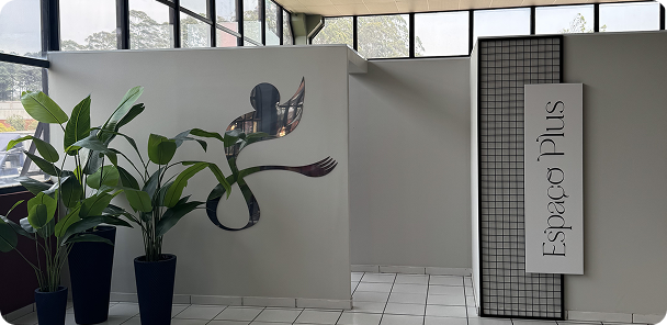 Espaço interno iluminado com plantas em vasos pretos à esquerda e uma parede branca com um logo decorativo prateado e o texto 'Espaço Plus' em painel na direita.