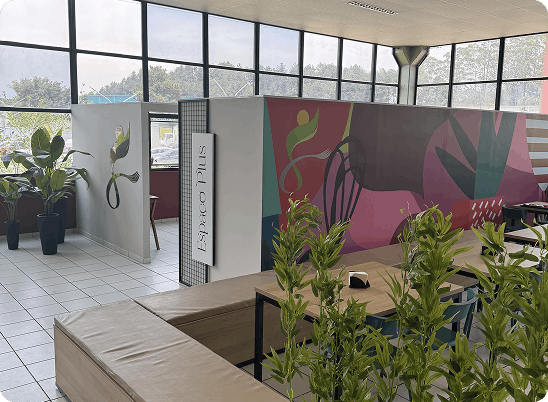 Interior de cafeteria com grandes janelas, plantas decorativas, assentos de madeira, e parede com arte abstrata colorida e texto Espaco Plus.