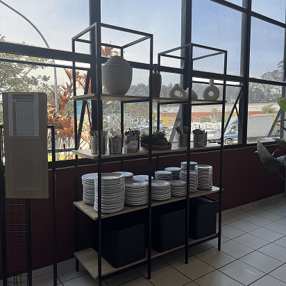 Estante preta com prateleiras de madeira contendo pratos empilhados, utensílios metálicos e vasos decorativos, encostada em janela grande com vista para área externa ensolarada.