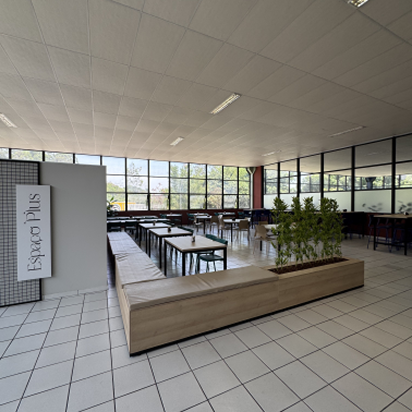 Área interna de cafeteria moderna com mesas, cadeiras, plantas em jardineira de madeira e grande parede de vidro com vista externa.