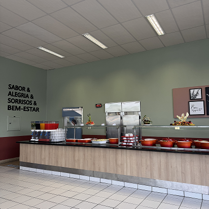 Buffet counter with various food containers, drinks, and plates in a clean dining area with green walls and motivational words in Portuguese on the wall.