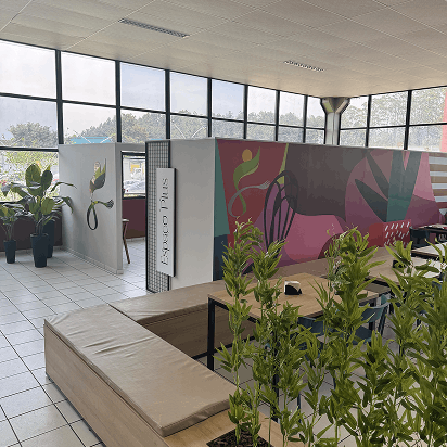 Área interna moderna com paredes de vidro, divisórias brancas, plantas em vasos e banco de madeira com almofada marrom em espaço aberto e bem iluminado.