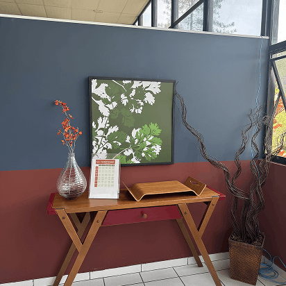 Mesa de madeira com vaso de vidro com flores secas, calendário branco e suporte de livro, contra parede meio azul e meio vermelha, quadro com folhas brancas e verdes ao centro e vaso com galhos secos ao lado direito.