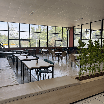 Interior de refeitório ou área de refeições com mesas, cadeiras, grandes janelas com vista para árvores e planta em vaso grande separando um espaço.