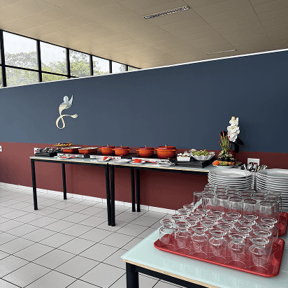 Mesa de buffet com várias travessas vermelhas, pratos, copos e decoração floral em ambiente moderno com parede azul e vermelha e luz natural por janelas amplas.