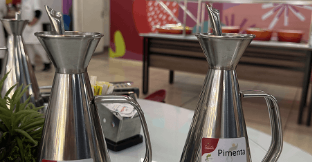 Dois recipientes de metal para temperos, um identificado como pimenta, sobre uma mesa branca com fundo desfocado de um ambiente interno.