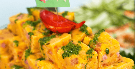 Cubos de prato amarelo com pedaços verdes e vermelhos, decorados com um molho verde e um pedaço de tomate no topo.