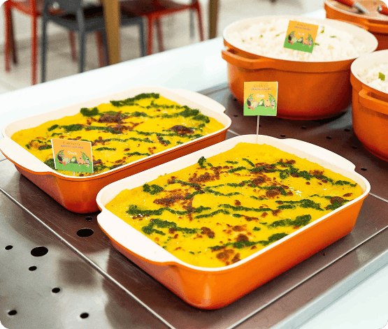 Duas travessas laranjas retangulares com um prato gratinado amarelo decorado com molho verde, em uma bancada metálica, com outras travessas ao fundo.