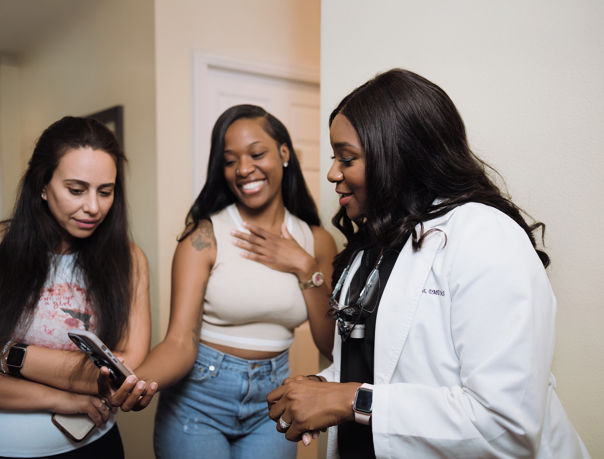 Three women, one in a white medical coat with a stethoscope, engaging in a friendly conversation indoors.