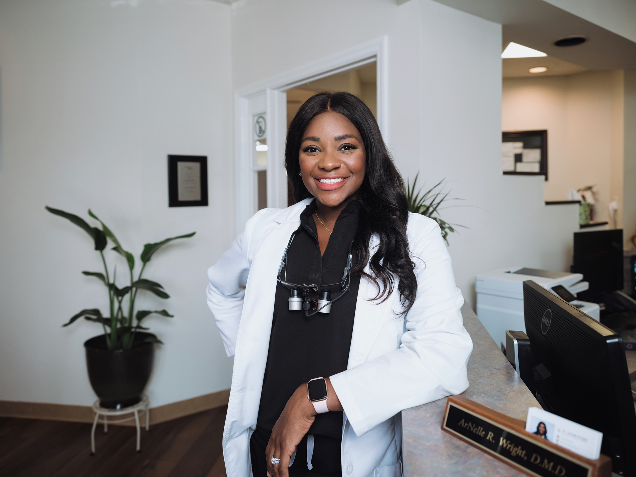 Smiling female dentist in a white coat with dental loupes around her neck leaning on a reception desk in a dental office.