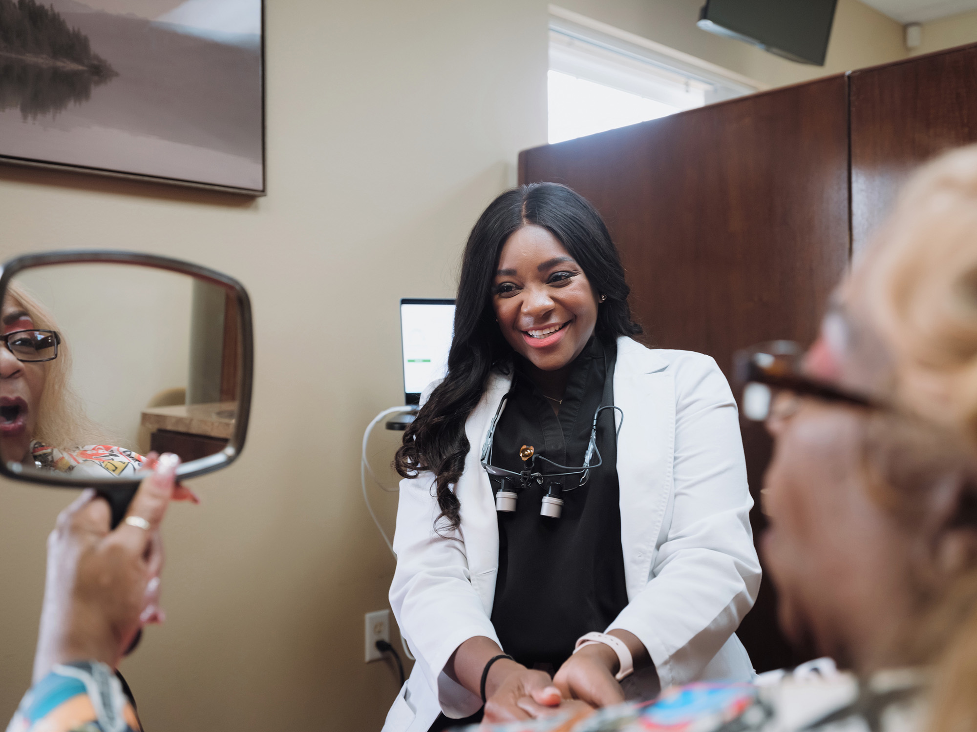 Female dentist in white coat smiling and holding hand of a patient who is looking into a handheld mirror.