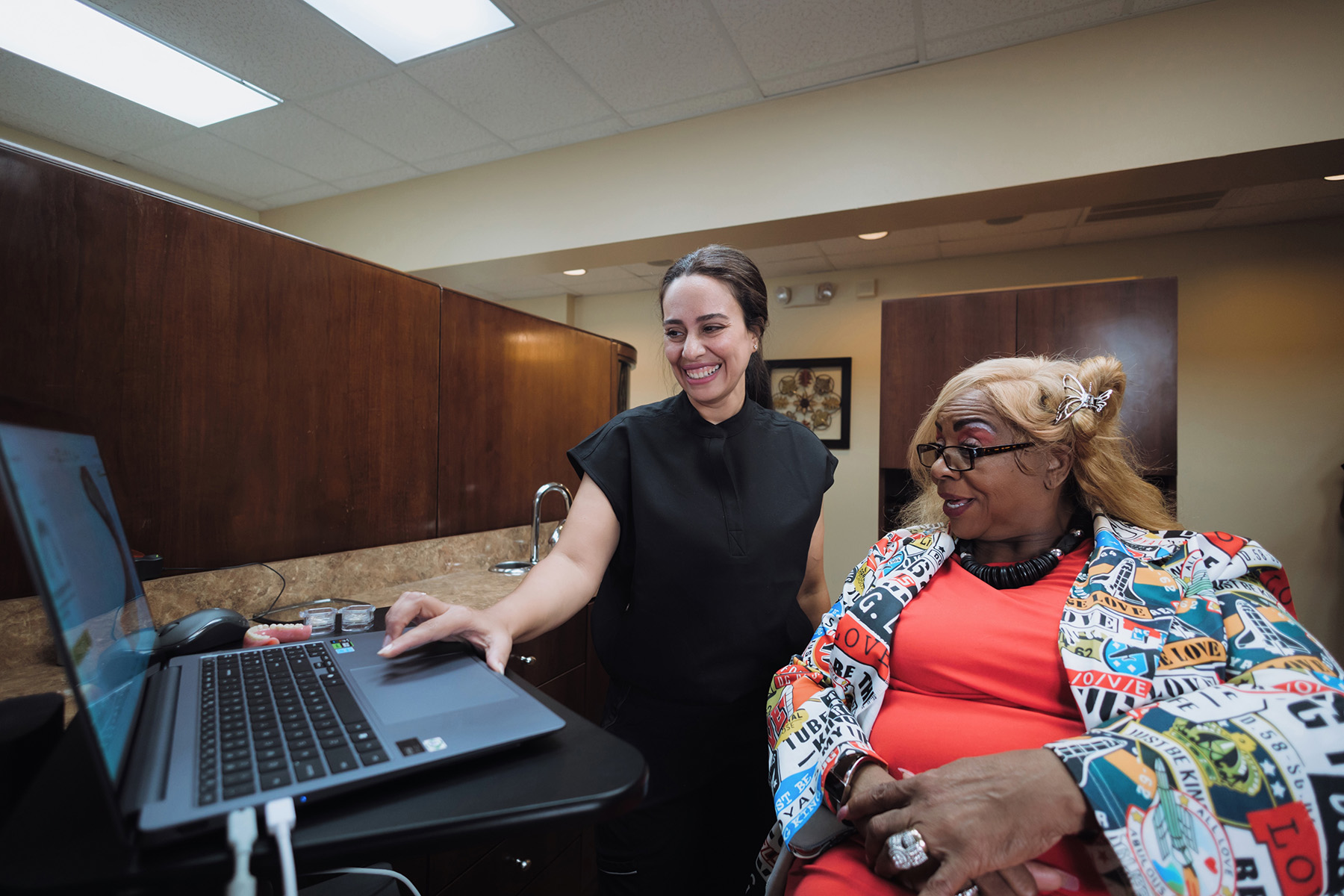 A smiling healthcare professional shows a laptop screen to a seated woman wearing glasses and a colorful jacket in a medical office.