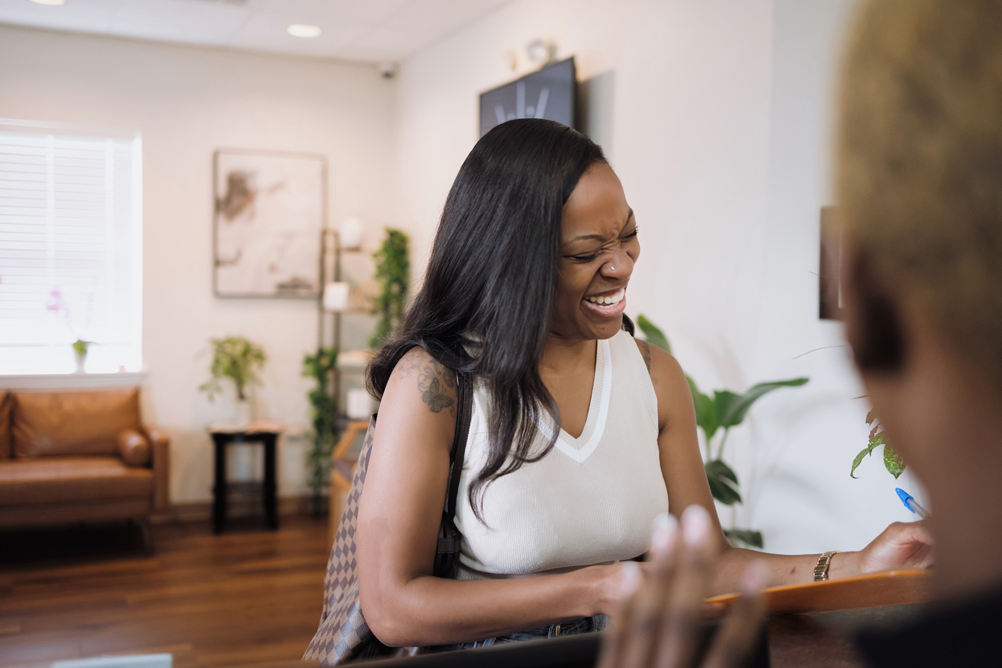 Woman smiling and laughing while interacting with a person at a reception desk in a cozy, well-lit room.