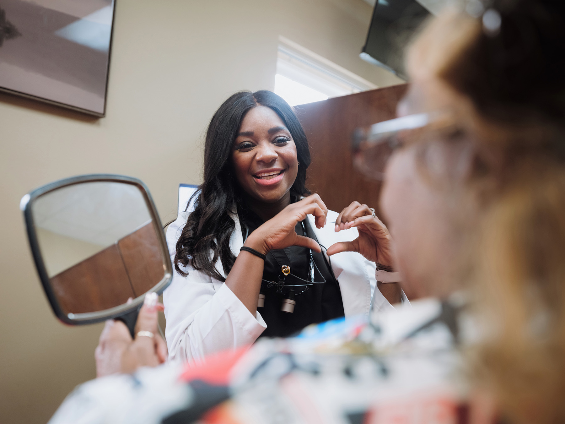 Smiling female healthcare professional making a heart shape with her hands to a patient holding a mirror.