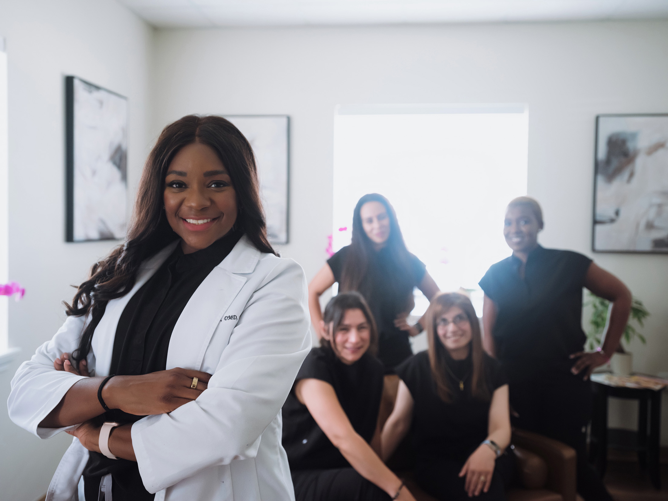 Confident female doctor in a white coat with arms crossed in front of a group of four women in black clothing in a well-lit office.