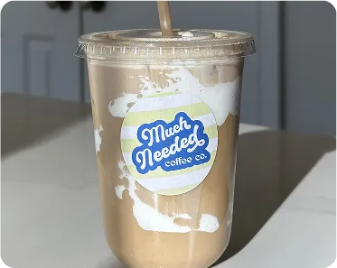 Plastic cup filled with iced coffee with a straw and a label that reads 'Much Needed coffee co.' on a table indoors.