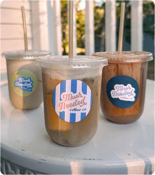 Three plastic cups of iced coffee with lids and straws on a round white table outdoors, each labeled with a circular blue and white striped sticker reading 'Much Needed coffee co.'