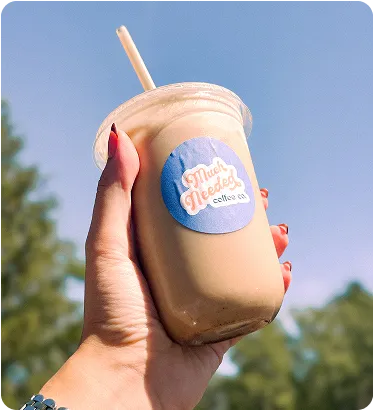 Hand holding a clear plastic cup with a straw containing a light brown iced coffee against a blue sky and blurred green trees background, labeled 'Much Needed coffee co.'