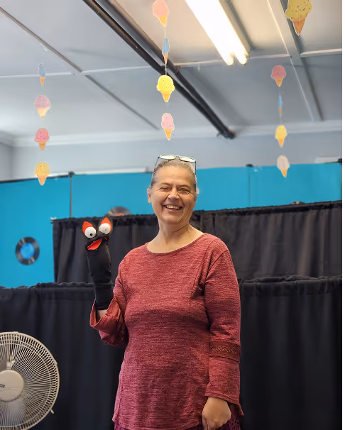Smiling woman in a maroon sweater holding a black sock puppet with orange eyes indoors with hanging ice cream decorations.
