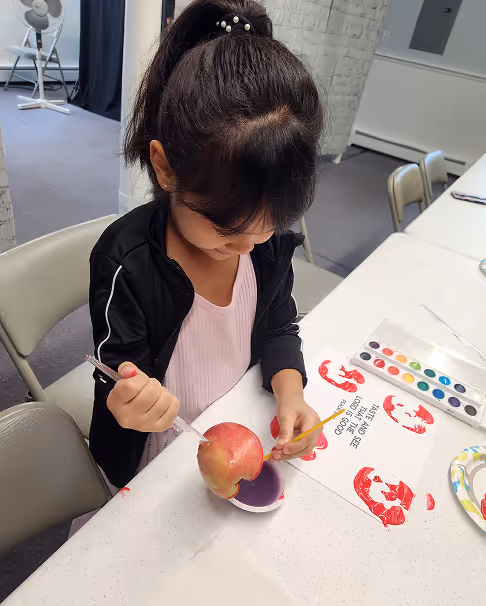 Young girl with a ponytail painting a red apple at a white table with watercolor paints and shrimp stamped paper nearby.